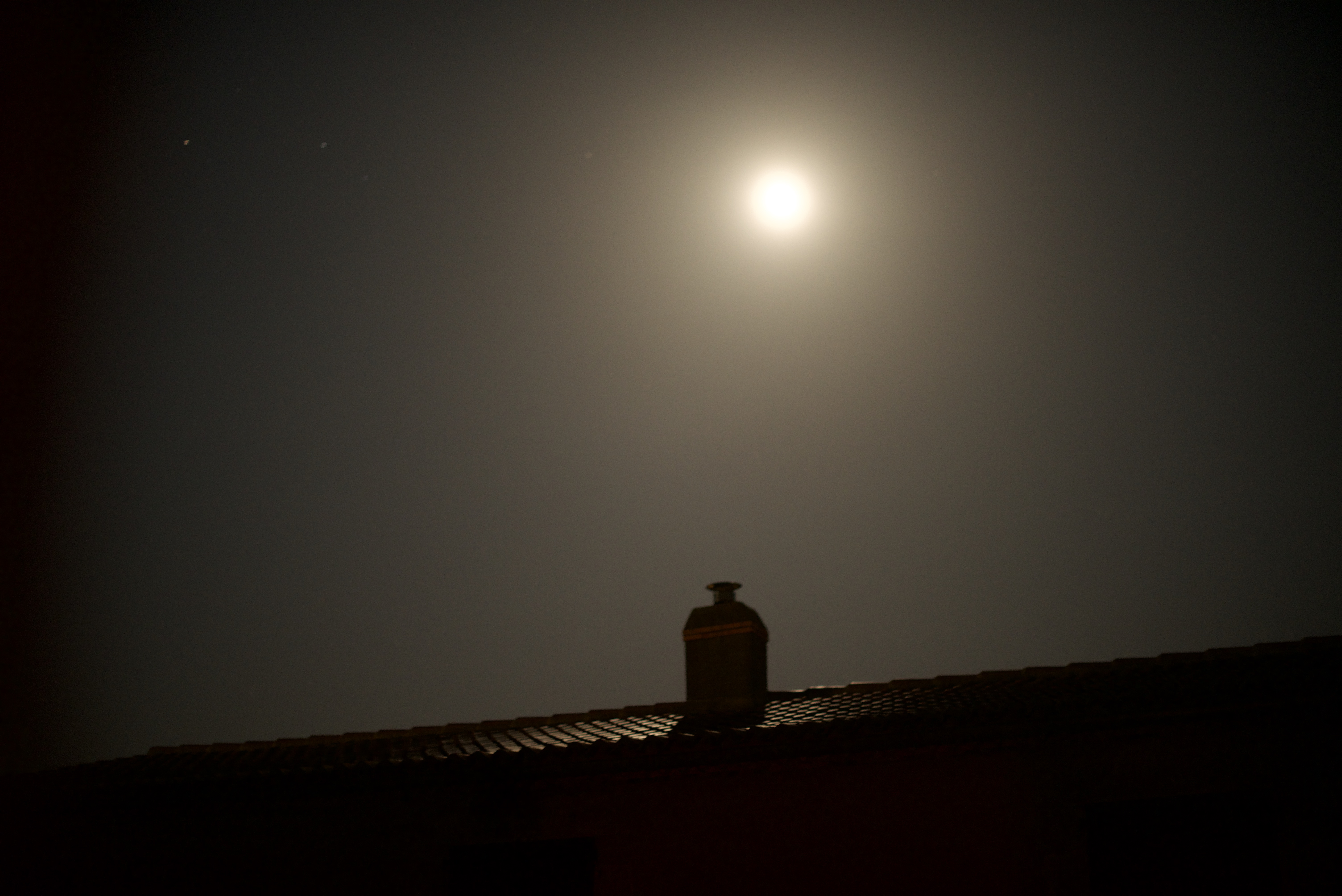Photo de la Lune, qui brille comme un soleil, au dessus d'un toit de tuiles.