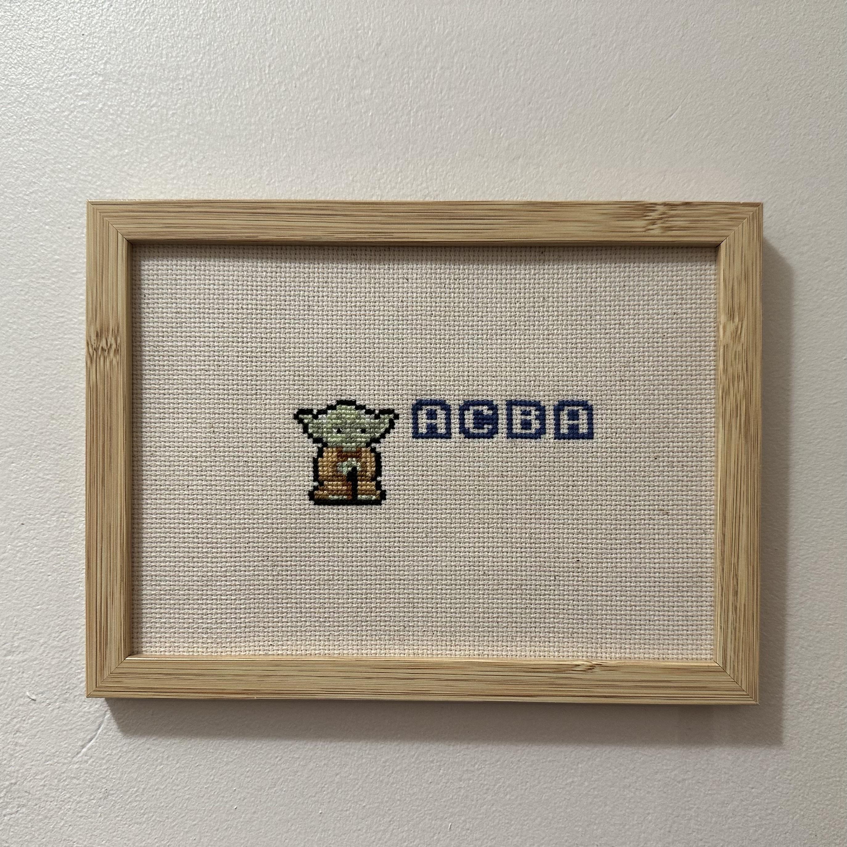 Yoda en pixel art avec les lettres ACBA dans une fonte de jeux videos.  La broderie est sur une toile écrue, dans un cadre en bois.