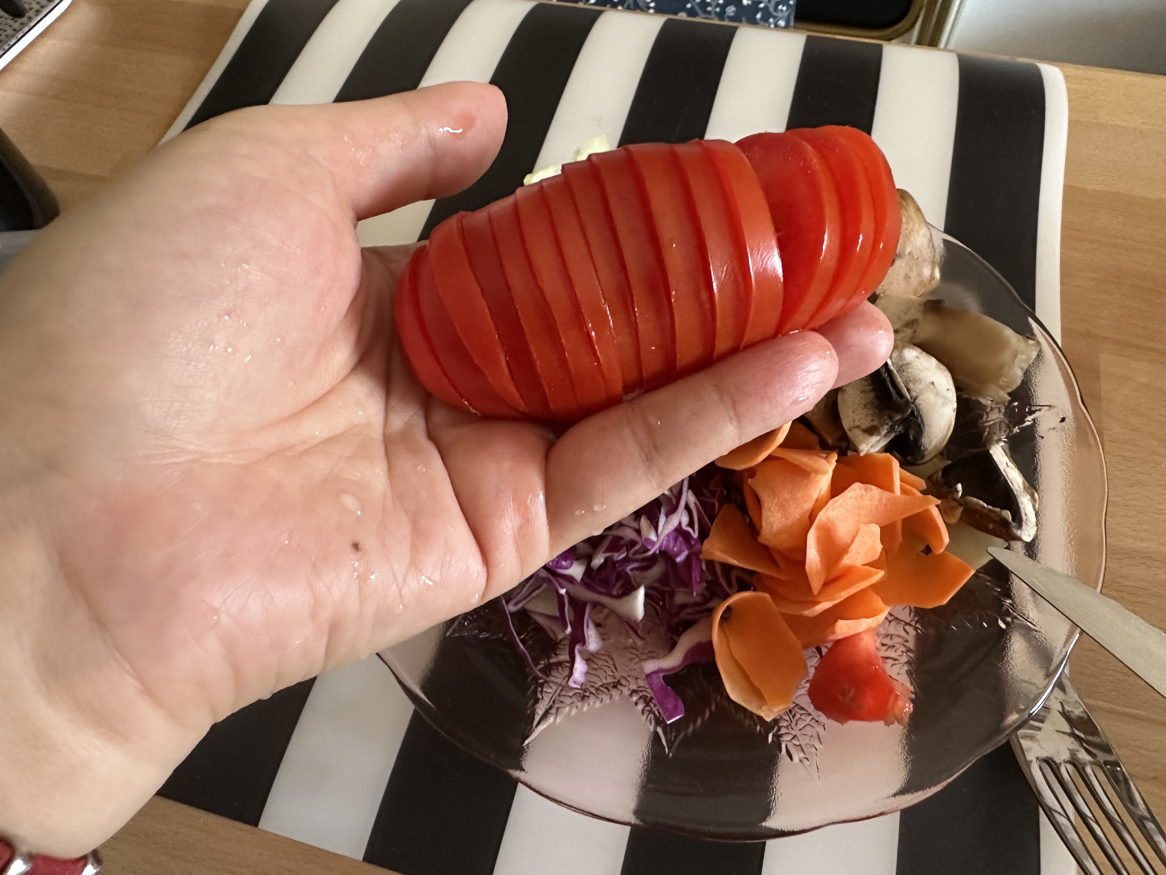 Une tomate coupée en tranches très fines, directement dans ma main gauche au dessus de mon assiette de salade, sur un set de table à larges rayures blanches et noires.