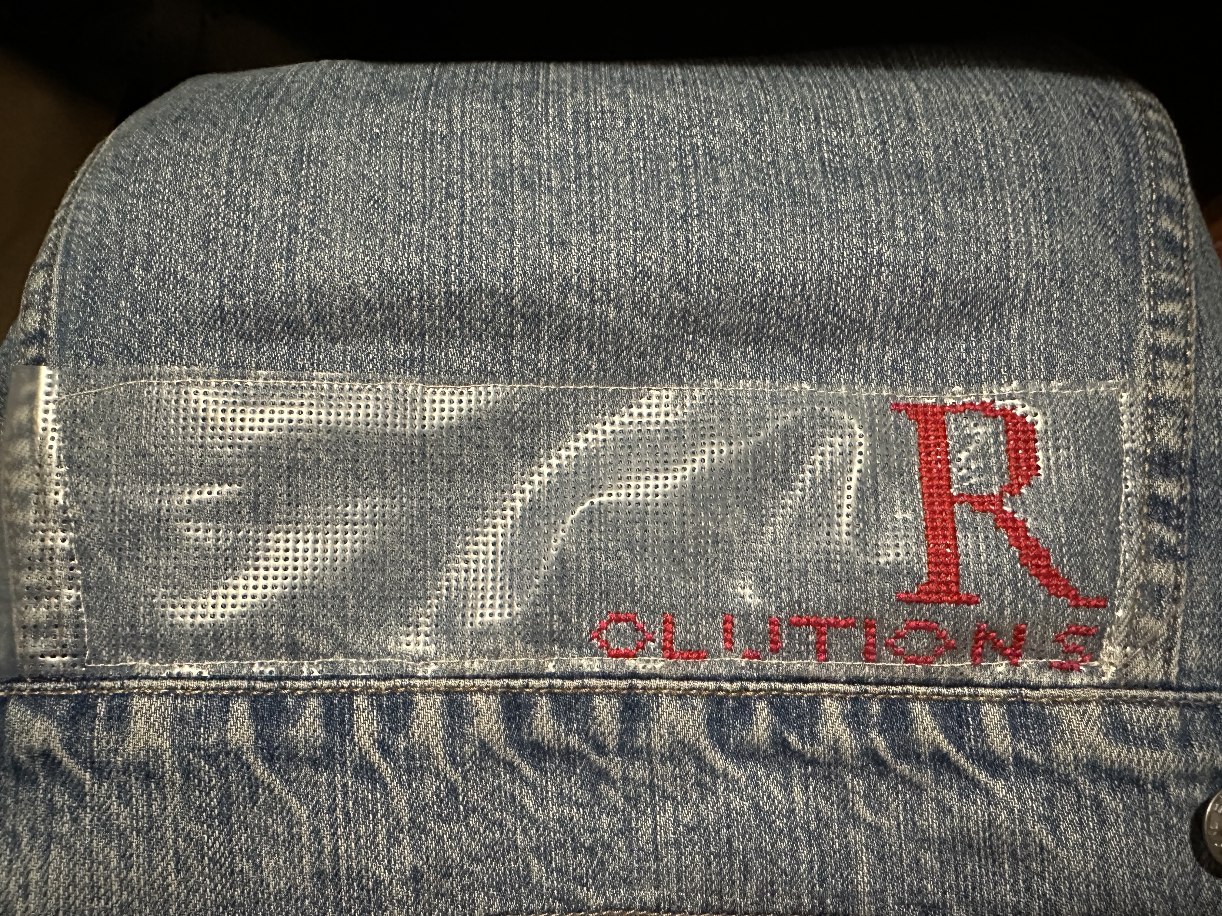Le bas du dos d'une veste en denim, une lettre R majuscule est brodée en rouge, et des lettres plus petites qui ne forment pas encore le mot SOLUTIONS