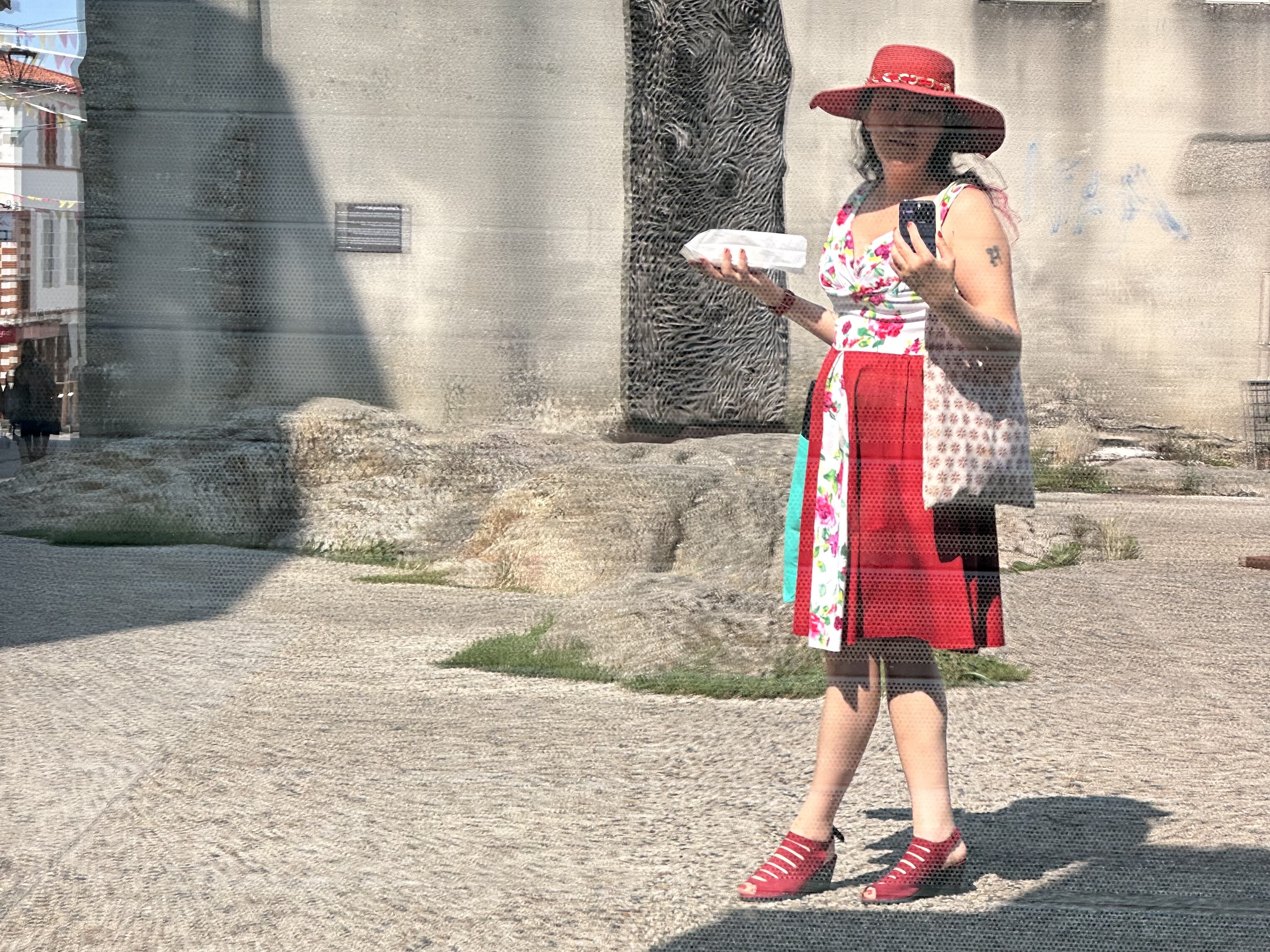 autoportrait dans une vitrine, je porte une robe rouge et blanche à fleurs, un chapeau rouge, un sac qui contient des bottes et un autre qui cache des patisseries. J'ai les jambes croisées au dessus de chaussures à talon rouge sombre.