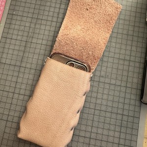 Pochette de cuir pile au format de la coque de mon iPhone 14 Pro.  Le cuir est rose. On voit cinq rivets sur chaque côté de la pochette, qui est posée à plat sur mon établi, avec un quadrillage centimétrique.